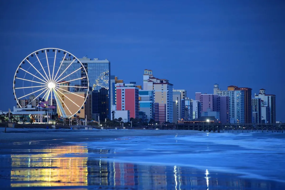 A cheerful waterfront scene featuring a Ferris wheel, colorful buildings, and their reflections on the water. The open-air, festive setting evokes the joy of exploring Myrtle Beach’s entertainment and dining options.