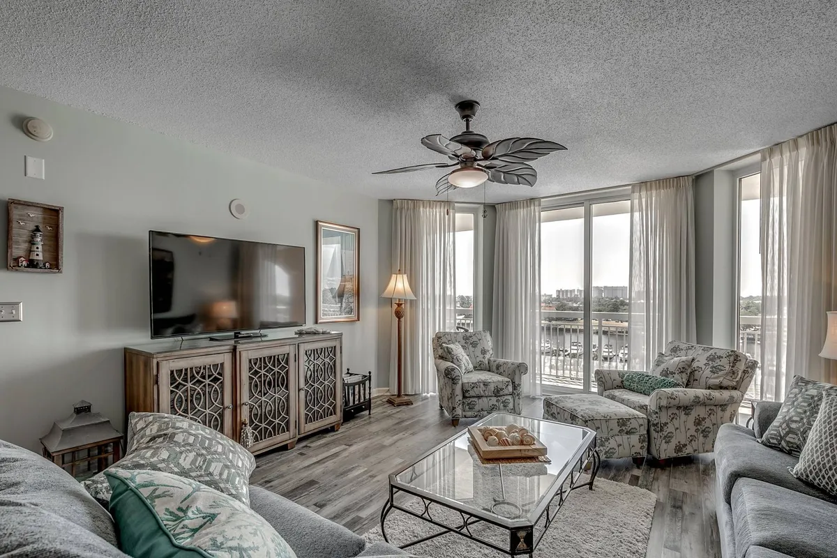 A comfortable seating area bathed in natural light from large windows, with a TV unit, decorative coffee table, and ample space for family to relax, socialize, or enjoy a movie night. The windows offer glimpses of scenic water views, connecting the interior with the outdoors.