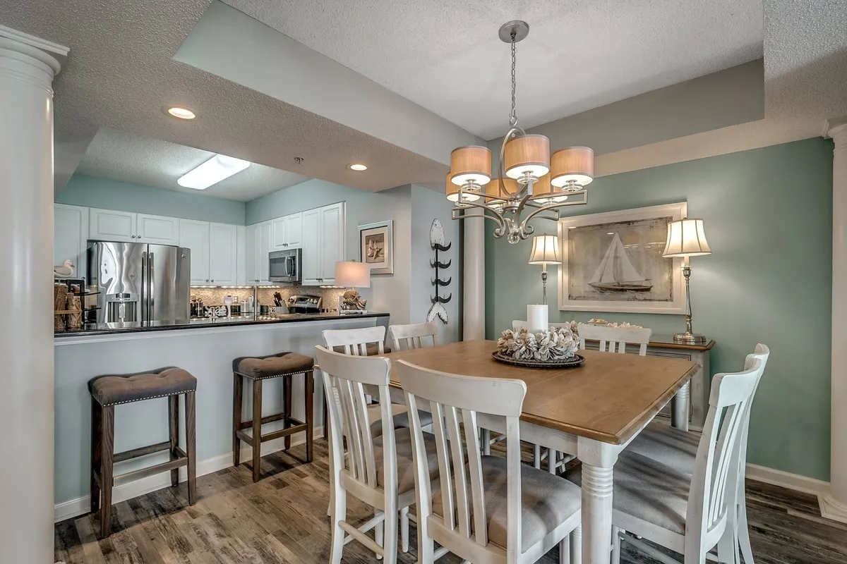 A spacious dining area with a large table set beneath a chandelier, adjacent to a kitchen counter. Wall art adds a decorative touch, and the area is brightly lit, ideal for family meals and social gatherings.