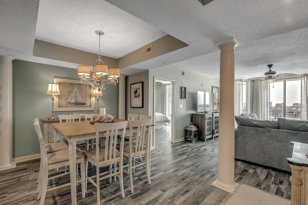 A spacious open-concept dining and living area, featuring a large dining table beneath a chandelier, comfortable seating, decorative pillars, and a view into the inviting living space. The scene is styled for social gatherings, with room for groups to relax, dine, and entertain together.