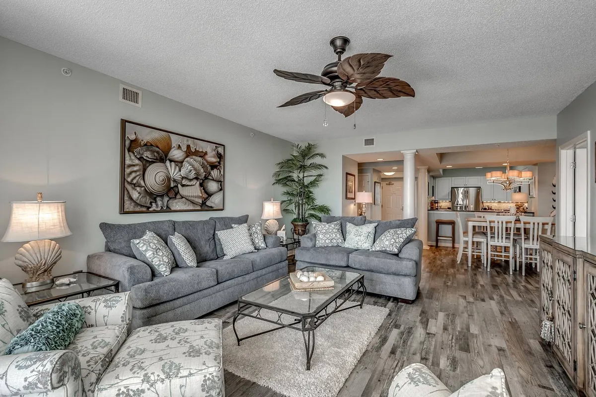 A welcoming living room featuring comfortable gray sofas adorned with decorative pillows, seashell-themed artwork on the wall, a glass coffee table, and a touch of greenery with a potted plant. The space is designed for relaxing with family, entertaining guests, or cozying up for a movie night.