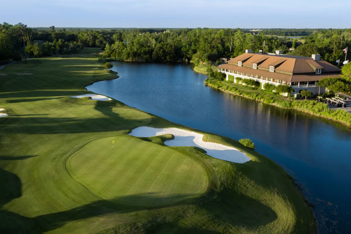 A serene view across a rolling green golf course, with sand traps, a picturesque lake, and a stately clubhouse in the background. The landscape is peaceful, inviting golf enthusiasts and those seeking relaxation amid scenic surroundings.