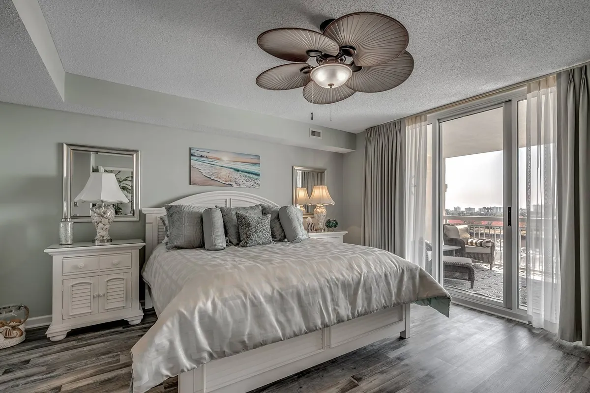 A serene king-sized bedroom with a large bed dressed in soft linens, a ceiling fan above, decorative artwork on the walls, and a sliding glass door that lets in abundant natural light. The door leads to a balcony, offering glimpses of water views and a tranquil setting, ideal for relaxation and waking up refreshed.