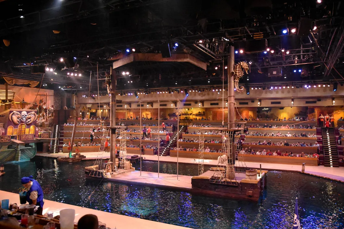 A dynamic interior scene of a live theatrical performance, featuring an elaborate pirate ship set, water stage, and audience seating. The immersive atmosphere suggests memorable entertainment and exclusive access to local shows and attractions.