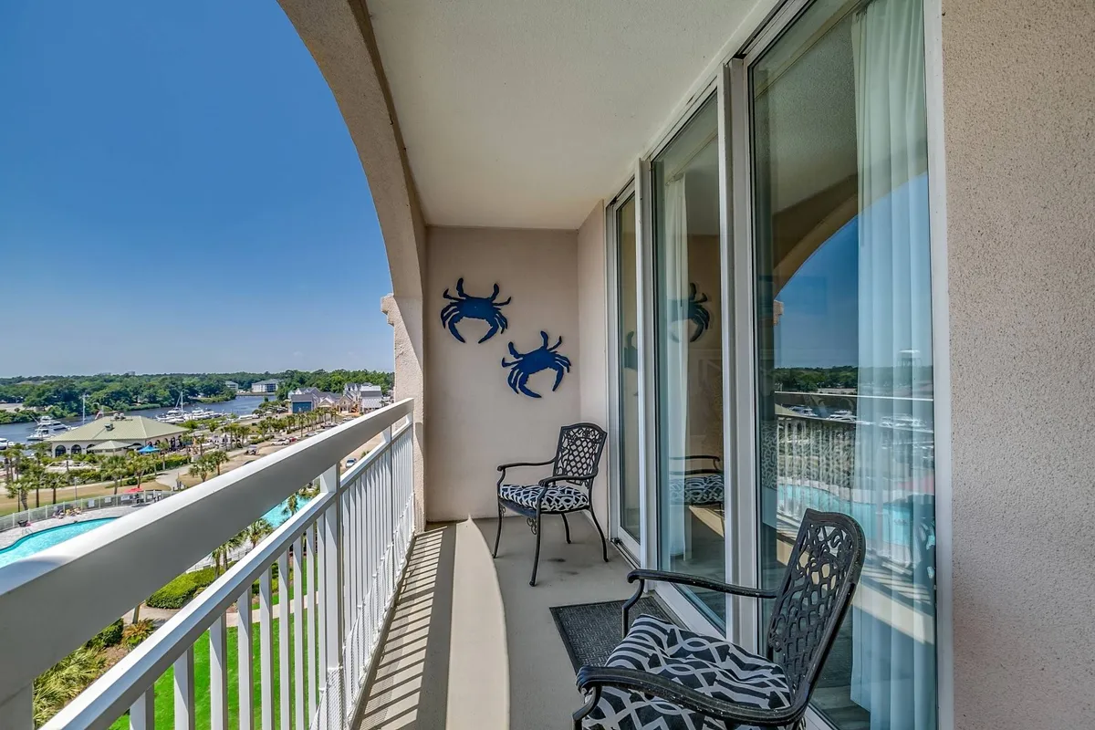 This image showcases the condo's private balcony with comfortable outdoor seating and distinctive decorative crab wall art. The balcony overlooks the stunning Intracoastal Waterway, with panoramic vistas stretching into the distance and capturing the sense of tranquility offered by the water views. The positioning allows for both sunrise and sunset enjoyment, and the setup encourages morning coffee or sunset cocktails outdoors.