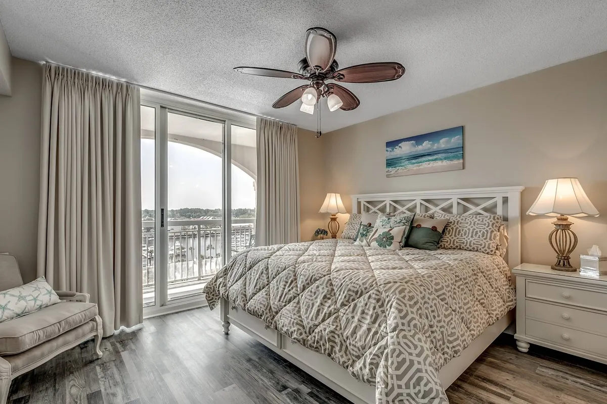 A cozy king bedroom featuring a large bed with decorative pillows, generous windows letting in soothing natural light, a ceiling fan, and nightstands with lamps. The setting is designed for relaxation and restful nights, with inviting tones and a peaceful ambiance.