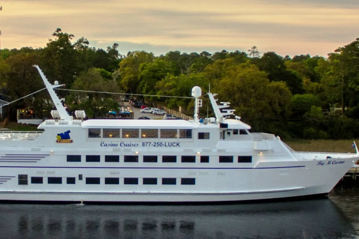 This exterior image shows a casino cruise ship docked along the waterfront, surrounded by lush trees and calm water. The vessel is lively and festive, suggesting opportunities for entertainment and socializing on the water, and providing a scenic view of the natural surroundings and sunset.