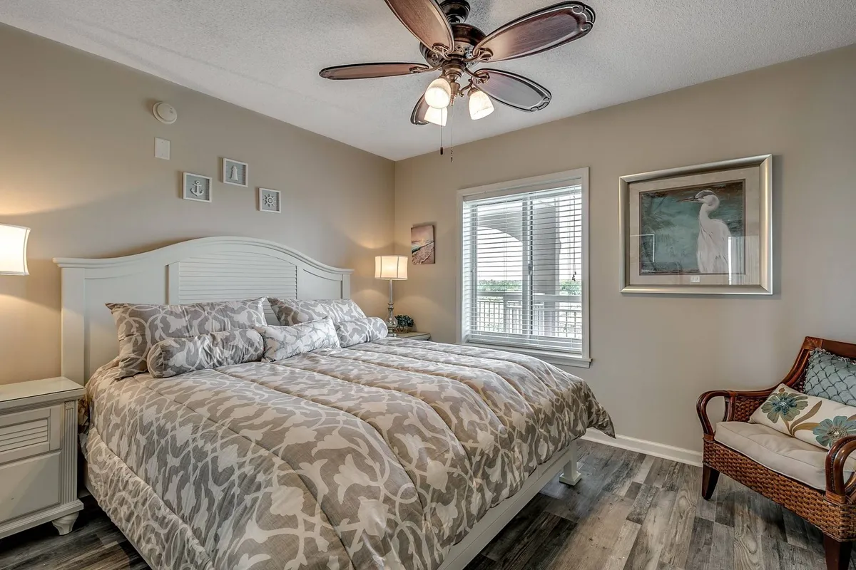 A calm, sunlit bedroom with a cozy bed, large window, tasteful artwork, and a comfortable chair. The natural light streaming in hints at a welcoming start to the day and offers a subtle connection to the outdoors.
